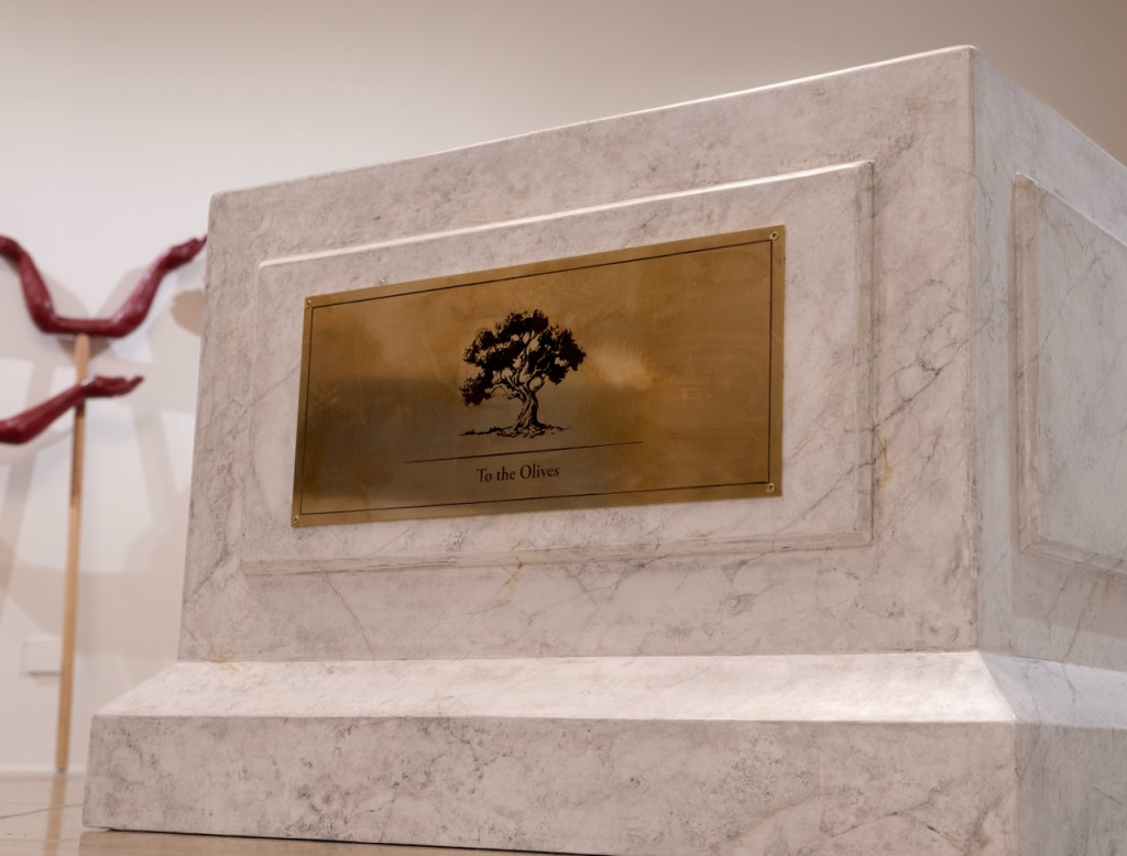Nadia Kaabi-Linke, To the Olives, 2026. Side view of the white marble plinth with a framed panel and engraved brass olive tree label in a gallery.