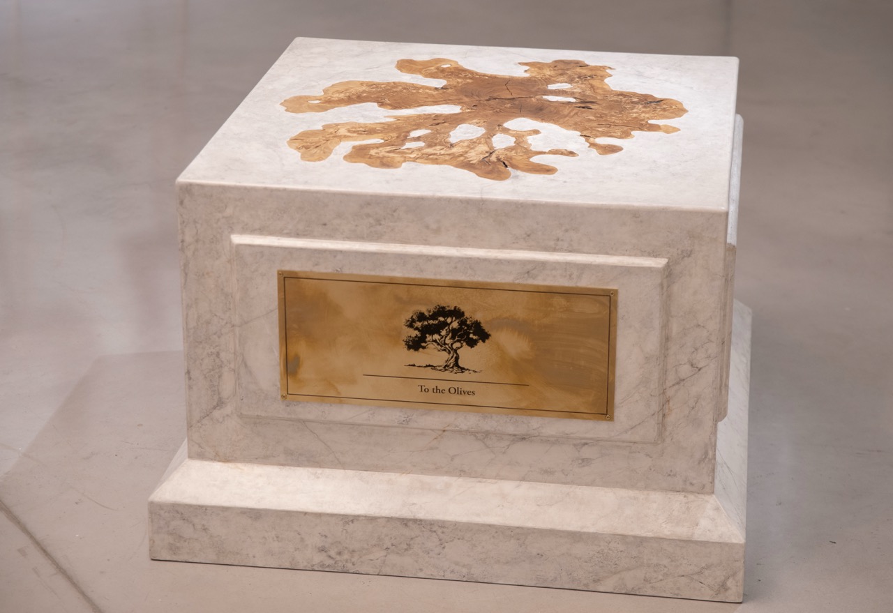 Nadia Kaabi-Linke, To the Olives, 2026. Front view of the marble plinth sculpture with an olive wood slice on top and a brass plaque engraved with an olive tree.